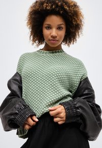 Fiatal nő göndör hajjal, texturált világoszöld pulóvert és túlméretezett fekete bőrkabátot visel, egyszínű háttér előtt.