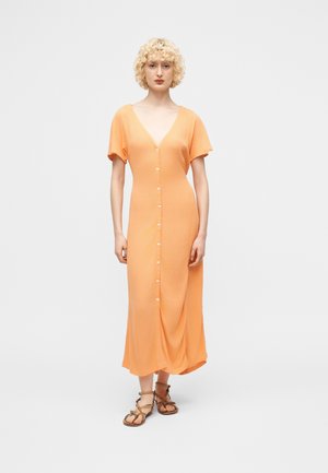 Femme aux cheveux blonds bouclés portant une robe midi boutonnée couleur pêche et des sandales plates marron à lanières, debout devant un fond uni.