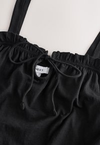 Črna majica brez rokavov z nabranim izrezom, zavezovanjem na vrvico in mehkim tekstilom. Na voljo je vidna etiketa z napisom blagovne znamke "NEXT".