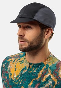 Gorra de ciclismo negra y gris con visera curvada, hecha de tela ligera. Presenta un diseño elegante y un detalle del logo en la parte frontal.