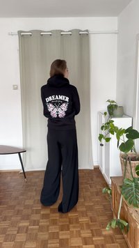 Črna kapuca z cvetličnim motivom in besedilom "DREAMER" na hrbtni strani, v kombinaciji z ohlapnimi črnimi hlačami; oblečena v sodobnem notranjem okolju.