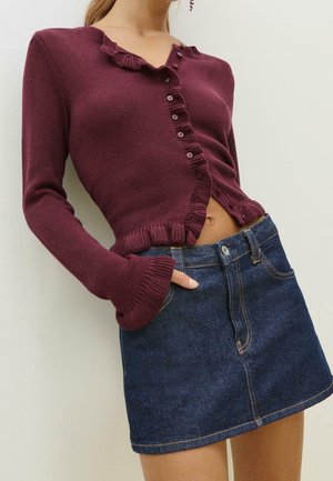 Femme portant un pull violet court à boutons avec des bords volantés et une mini-jupe en jean bleu foncé, main dans la poche.