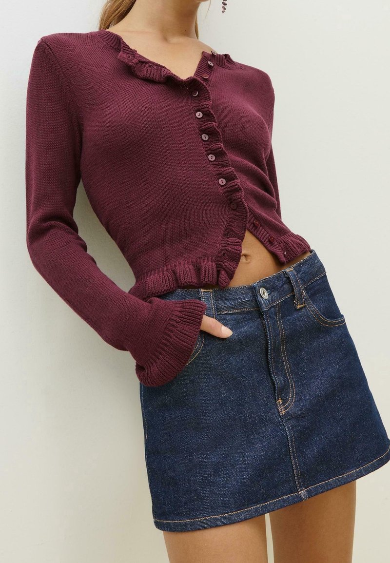 Femme portant un pull violet court à boutons avec des bords volantés et une mini-jupe en jean bleu foncé, main dans la poche.