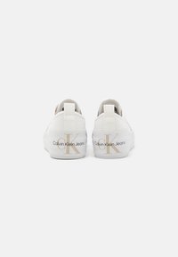 Sneaker bianche in tela con suola piatta, dotate di un logo dorato "Calvin Klein Jeans" e di un accento "CK" ben visibile sul tallone.