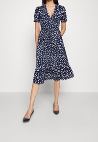 Tummansininen kietaisumekko, jossa on valkoista kukkakuviota, lyhyet puffihihat, solmittava vyötärö ja röyhelöhelma. Tummat stiletot täydentävät asua.