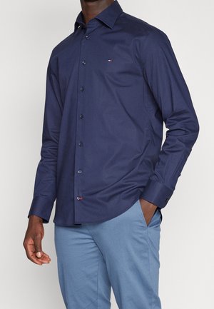 Homme portant une chemise bleu marine boutonnée à manches longues et un pantalon bleu clair, main dans la poche, sur un fond uni.