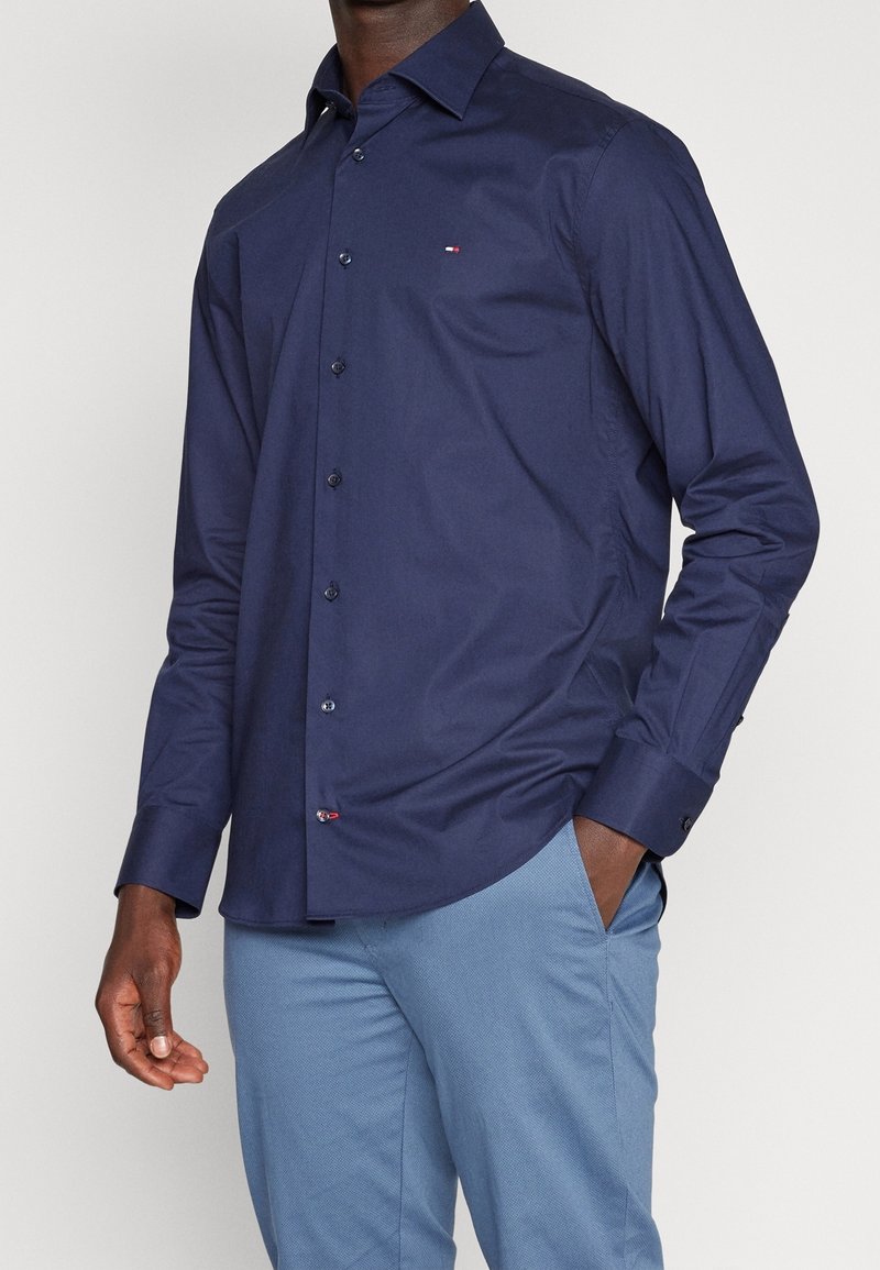 Homme portant une chemise bleu marine boutonnée à manches longues et un pantalon bleu clair, main dans la poche, sur un fond uni.