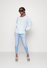 Camisa de botão de manga longa azul clara; combinada com jeans skinny azul clara e mules de salto verdes; óculos de sol pretos completam o visual.