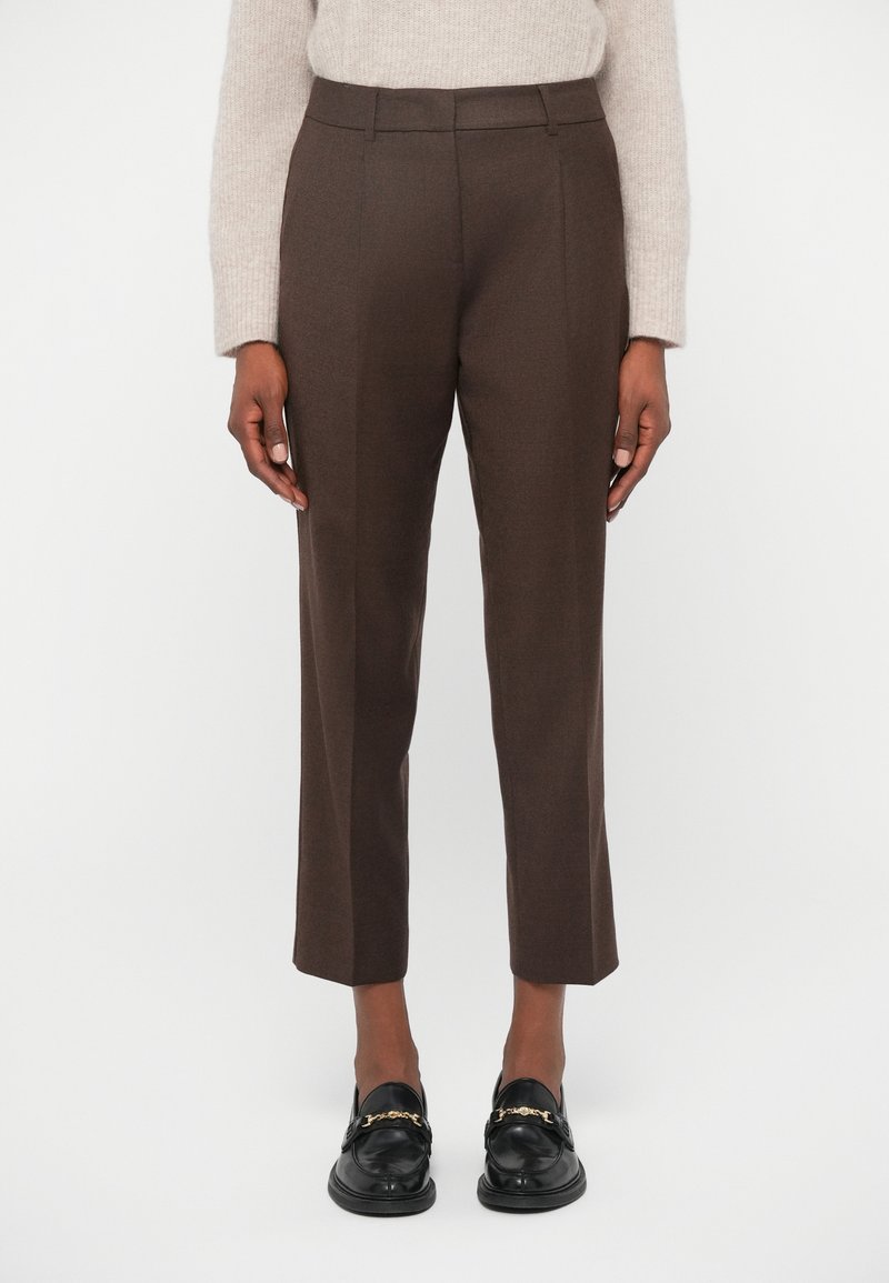 Καφέ παντελόνια με ίσια γραμμή, με απαλή υφή και διακριτές πιέτες, συνδυασμένα με μαύρες loafers που έχουν χρυσές λεπτομέρειες.