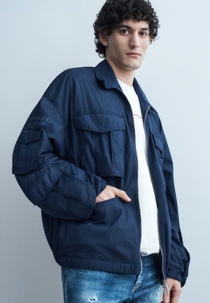 Jeune homme aux cheveux bouclés portant une veste bleu marine sur une chemise blanche et un jean bleu déchiré, debout devant un fond gris.