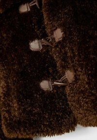 Brun luddig kappa med en texturerad, plyschaktig yta och bruna läderknäppningar. Kanterna är mjuka med en något oregelbunden finish.