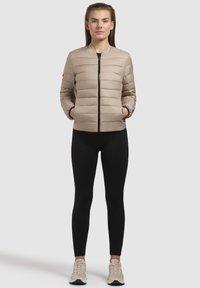 Giacca bomber imbottita beige con zip sul davanti, colletto a coste e tasche laterali, indossata sopra leggings neri e sneakers beige.