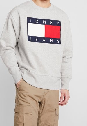 Mand iført lysegrå sweatshirt med stort rødt, hvidt og sort Tommy Jeans logo og beige cargobukser, hånd i lommen.