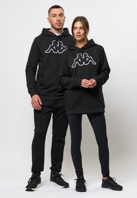 Zwarte hoodies met een wit Kappa-logo op de voorkant, gecombineerd met zwarte leggings en bijpassende sneakers. Glad materiaal, comfortabele pasvorm.