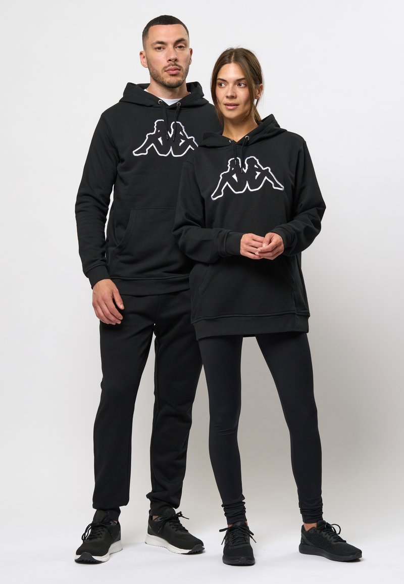 Zwarte hoodies met een wit Kappa-logo op de voorkant, gecombineerd met zwarte leggings en bijpassende sneakers. Glad materiaal, comfortabele pasvorm.