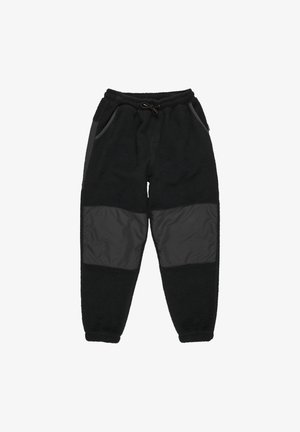 Garçon portant un bonnet orange, un t-shirt à manches longues sombre avec un logo, un pantalon polaire noir et des baskets New Balance marron, debout les mains dans les poches.