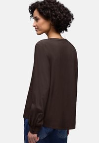 Blouse marron foncé à manches longues avec une coupe décontractée, texture lisse, encolure ronde et poignets à boutons, associée à un jean en denim.