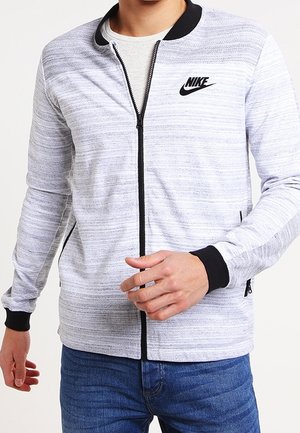 Mężczyzna w białej i szarej pasiastym dresie Nike zapinanym na zamek z czarnym kołnierzem i mankietami, w połączeniu z niebieskimi jeansami.