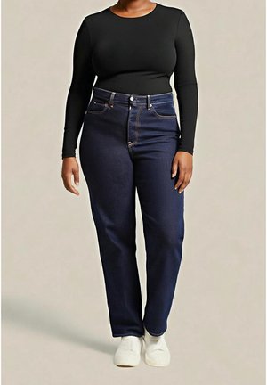 Person, die dunkelblaue High-Waist-Jeans, ein schwarzes Langarmshirt und weiße Sneakers trägt, steht vor einem schlichten beigen Hintergrund.