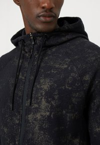 Sudadera oscura con material de forro polar texturizado y un sutil patrón de diseño, cierre de cremallera, capucha ajustable con cordón y acentos negros.