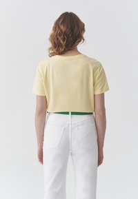 Camiseta amarilla recortada de algodón suave con mangas cortas, combinada con jeans blancos de cintura alta. Cinturón verde visible que acentúa.