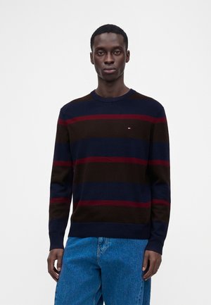 Homme portant un pull foncé avec des rayures horizontales rouges et bleu marine et un jean bleu, debout devant un fond blanc uni.