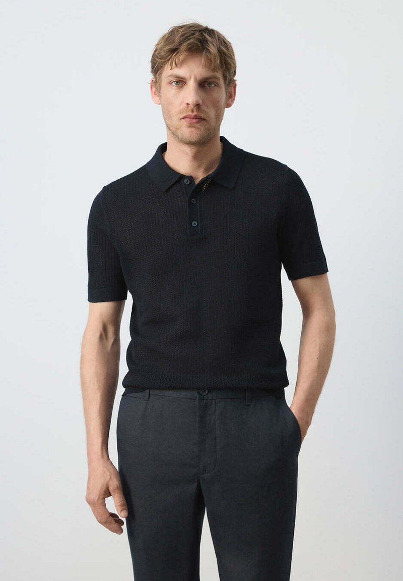Homme aux cheveux blonds courts, portant un polo en maille noir à manches courtes et un pantalon gris foncé, debout devant un fond clair et uni.