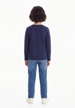 Enfant aux cheveux courts et bouclés, debout de dos, portant un t-shirt à manches longues bleu marine, un jean bleu et des baskets blanches sur fond blanc.
