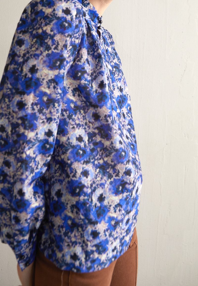 Personne portant un chemisier à manches longues avec un motif floral bleu et noir et un pantalon marron, debout devant un mur clair uni.
