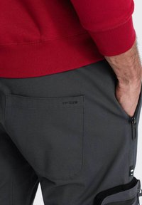 Graue Hose mit einer Gesäßtasche mit Etikett, strapazierfähigem Stoff und einer seitlichen Reißverschlusstasche, kombiniert mit einem roten Pulloverbund.
