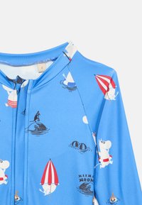 Modro oblačilo z zadrgo, na katerem so igrivi beli risani liki, ki držijo rdeče-bele dežnike in plavajo z življenskimi obročki.