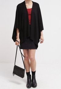 Persoon in zwarte gedrapeerde cardigan, rode top, zwarte rok, zwarte enkellaarsjes en met een zwarte schoudertas tegen een effen achtergrond.