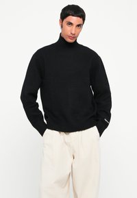 Suéter negro de cuello alto hecho de un tejido texturizado, con puños y dobladillo acanalados, combinado con pantalones cremosos de corte holgado.