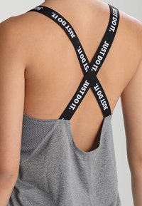 Grå atletisk tanktop med et mesh-design, der har sorte krydsede stropper mærket "JUST DO IT." Ribbet tekstur på stoffet.