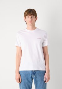 Hvit bomulls t-skjorte med rund hals, korte ermer, og en liten rød "Calvin Klein" logo på venstre bryst. Brukt sammen med blå denim jeans.