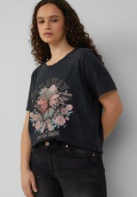 Zwarte T-shirt met korte mouwen in een vervaagde look; heeft een grafisch ontwerp van een vlinder en bloemelementen. Tekst luidt "VLEUGEL OP in de Nacht."