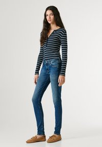 Naine seisab valge taustaga ees, kandes sinise ja valge triibulise pika varrukaga t-särki, siniseid kitsaid teksaseid ning beeže mokasäärista stiilis jalanõusid.