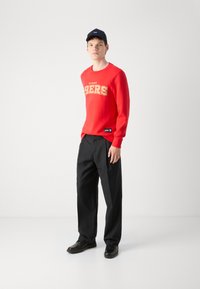 Sweatshirt vermelho com "49ERS" em dourado, calças pretas e ténis pretos. O sweatshirt tem um decote redondo e punhos canelados. Boné preto.