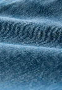 Detailní záběr na texturovanou modrou džínovinu s viditelným vzorem tkaní a jemnými záhyby, které vytvářejí jemné stíny.
