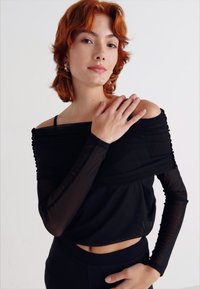 Μαύρη τοπ χωρίς ώμους με μακριές διαφανείς μανίκες, διαθέτοντας ριχτό σχέδιο και εφαρμοστό μέσο, που αναδεικνύει μαλακή υφή υφάσματος.