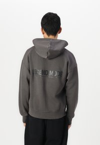 Sweat à capuche gris avec une coupe décontractée, présentant un imprimé réfléchissant au dos avec la mention "MODE LÉGENDE". Texture lisse, style décontracté.
