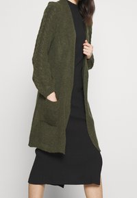 Olivgrön stickad cardigan med texturerade ärmar, öppen front och två framfickor, bärs över en svart ribbad klänning.