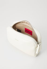 White leather cosmetic bag with silver zipper, featuring a beige interior lining and a red "HUGO" label inside.