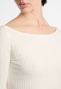 Maglietta bianca a maniche lunghe con scollo a barca. La texture morbida e il design aderente mettono in risalto la forma del corpo con sottili linee orizzontali.