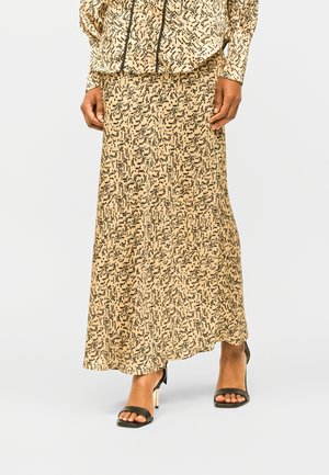 Beige Maxirock mit abstraktem schwarzem Muster, getragen mit passendem Langarmoberteil und schwarzen Sandalen mit Absatz und goldenen Details.