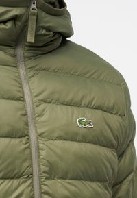 Veste matelassée vert olive avec fermeture éclair et petit logo crocodile vert brodé sur la poitrine près de la fermeture éclair.