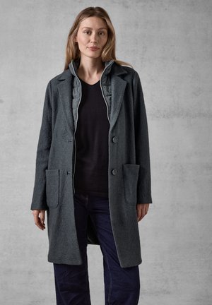 Cappotto in misto lana grigio con finitura testurizzata, rever a punta, due tasche frontali e chiusura a bottone, indossato sopra un top scuro.