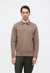 Pullover castanho com fecho zip e gola, textura ribateada e mangas compridas. Usado com calças verde-escuras. Design minimalista.