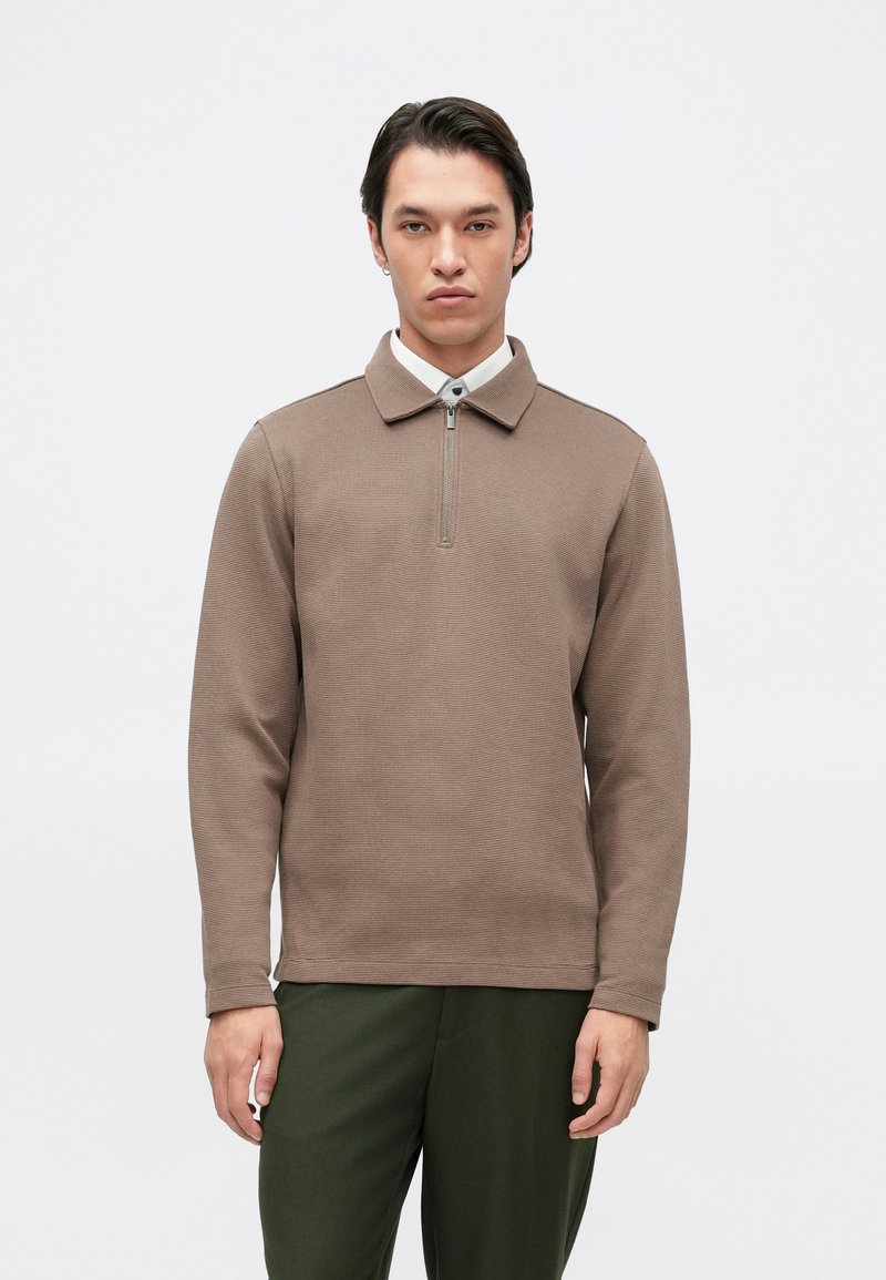 Pullover castanho com fecho zip e gola, textura ribateada e mangas compridas. Usado com calças verde-escuras. Design minimalista.