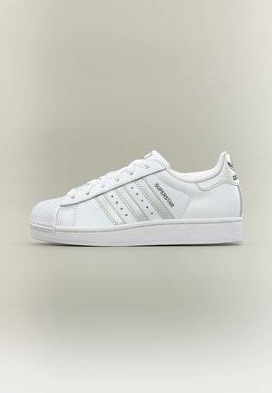 Baskets blanches Adidas Superstar avec trois bandes, lacets, bout en coque et marquage sur le côté et le talon sur un fond neutre.